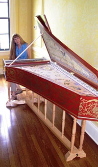 A white woman with long medium brown hair wearing a blue shirt is seated at a harpsichord. The harpsichord is red with gold arabesque decoration and is supported on a wooden stand.